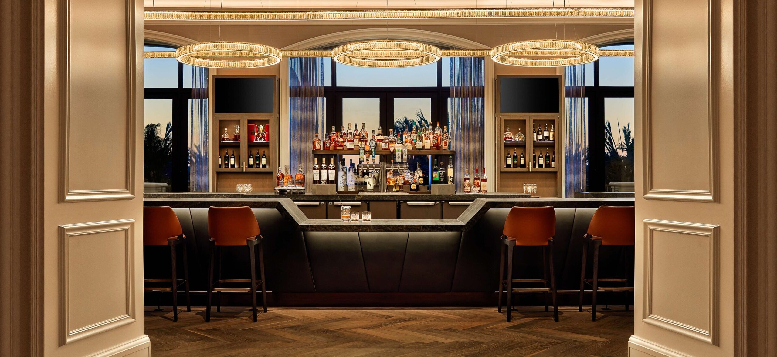 Upscale bar with leather seating, three circular chandeliers, and a well-stocked RealFood counter, viewed through double doors. Four barstools are positioned in front of the bar.
