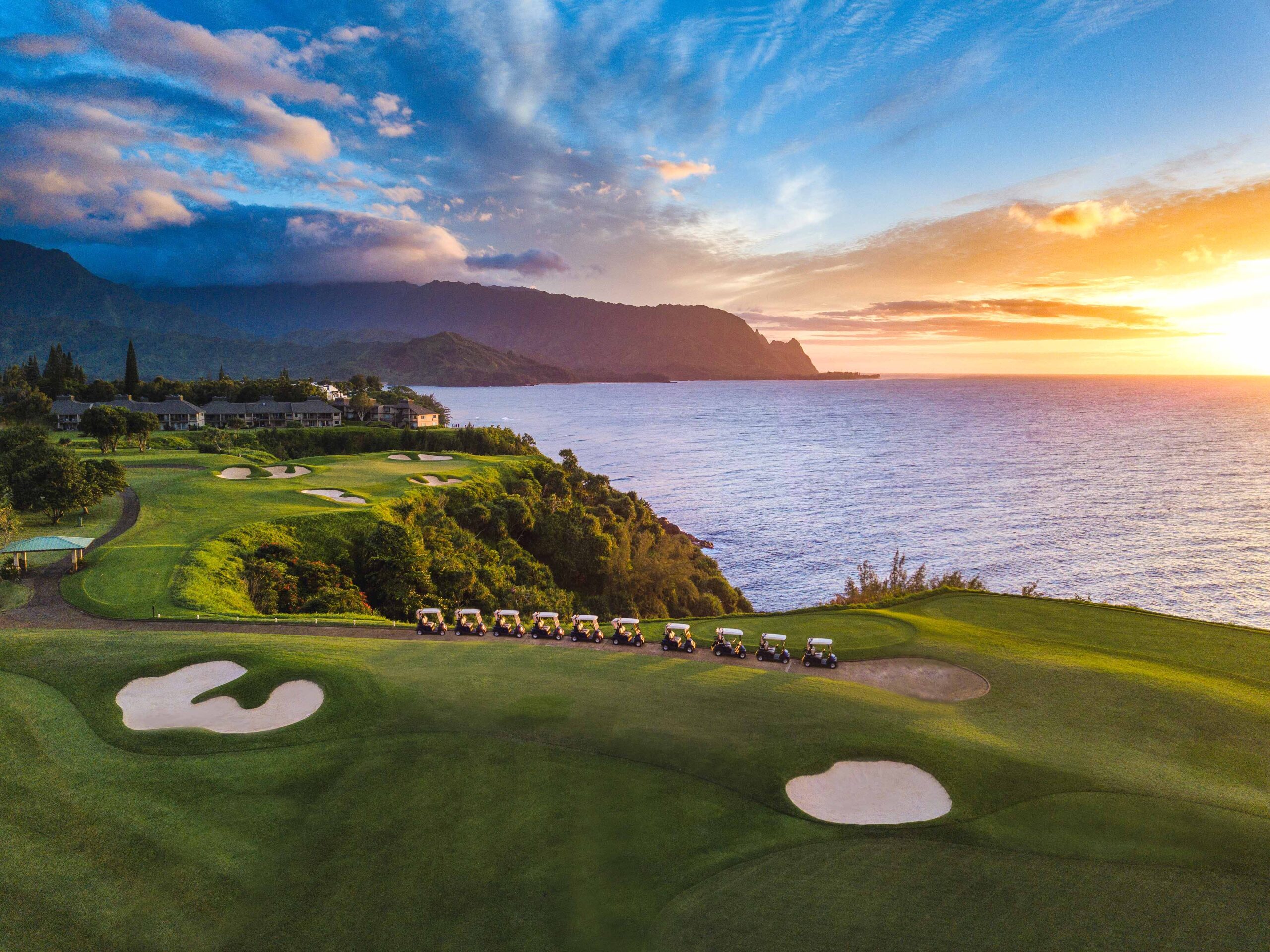 Golf course overlooking the ocean at sunset, with several golf carts lined up on the fairway and mountains in the background—perfect for relaxing after a round and enjoying RealFood with breathtaking views.