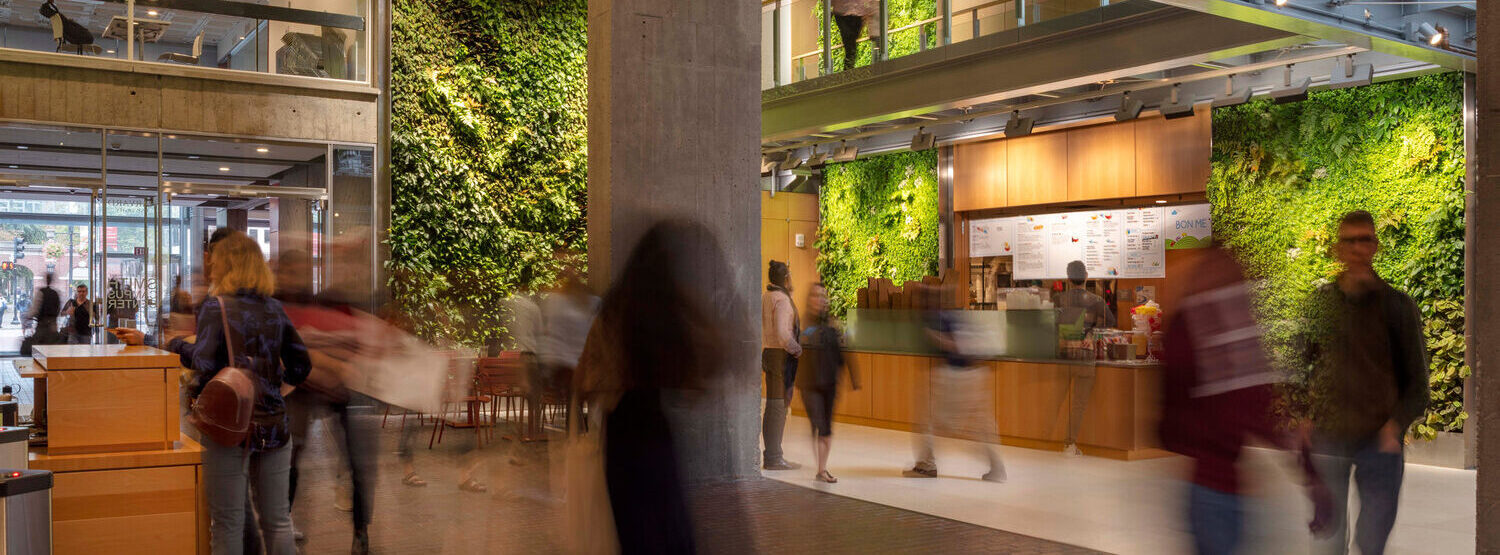 Modern indoor space with people walking, a RealFood cafe counter, glass walkways, and green living plant walls on concrete surfaces.