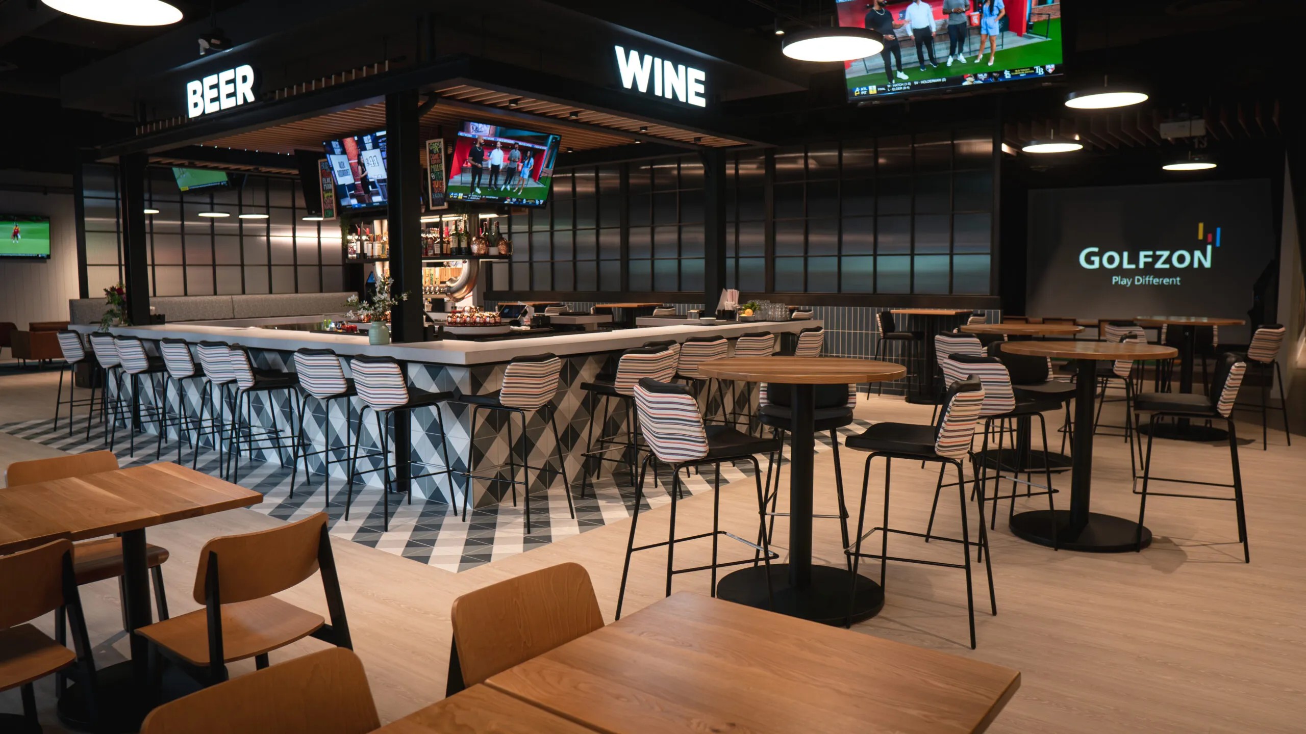 Modern bar area by RealFood with geometric-tiled counter, high-top tables, wooden chairs, and TVs displaying sports; illuminated 
