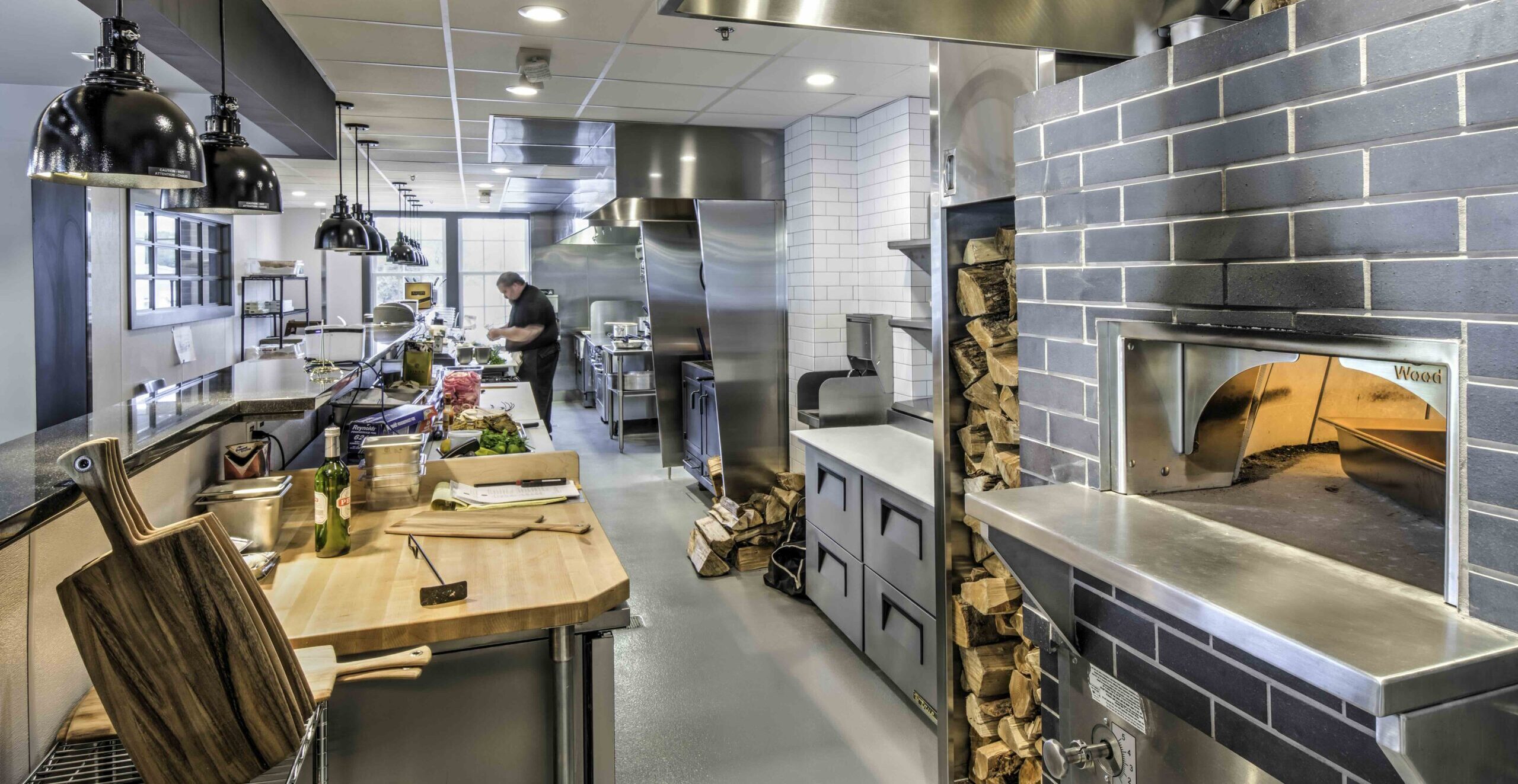Modern commercial kitchen with a wood-fired oven, food preparation area, stainless steel equipment, and a RealFood chef working in the background.