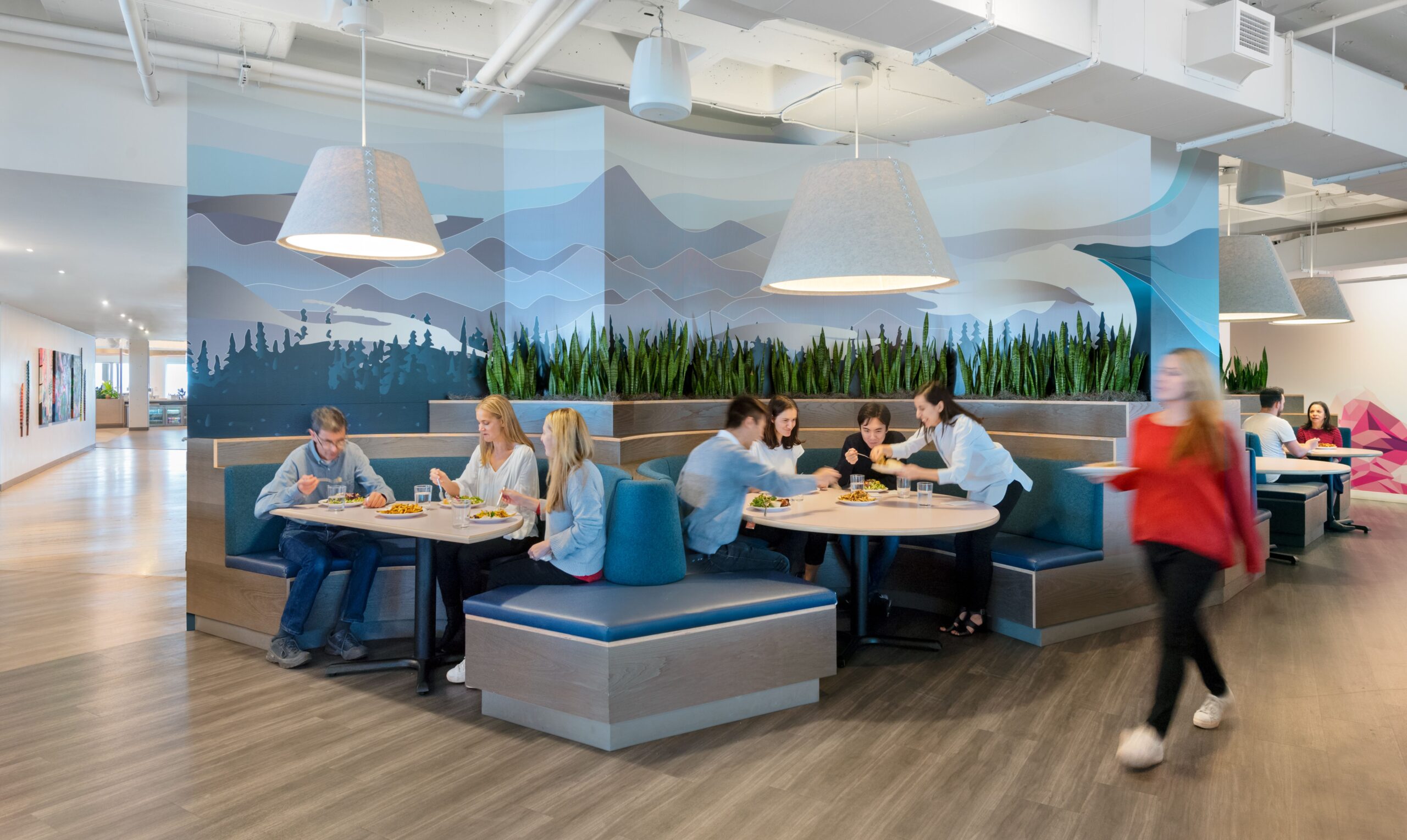 People enjoy RealFood at booth tables in a modern cafeteria with a mountain mural, while a person in a red top walks by in the foreground.