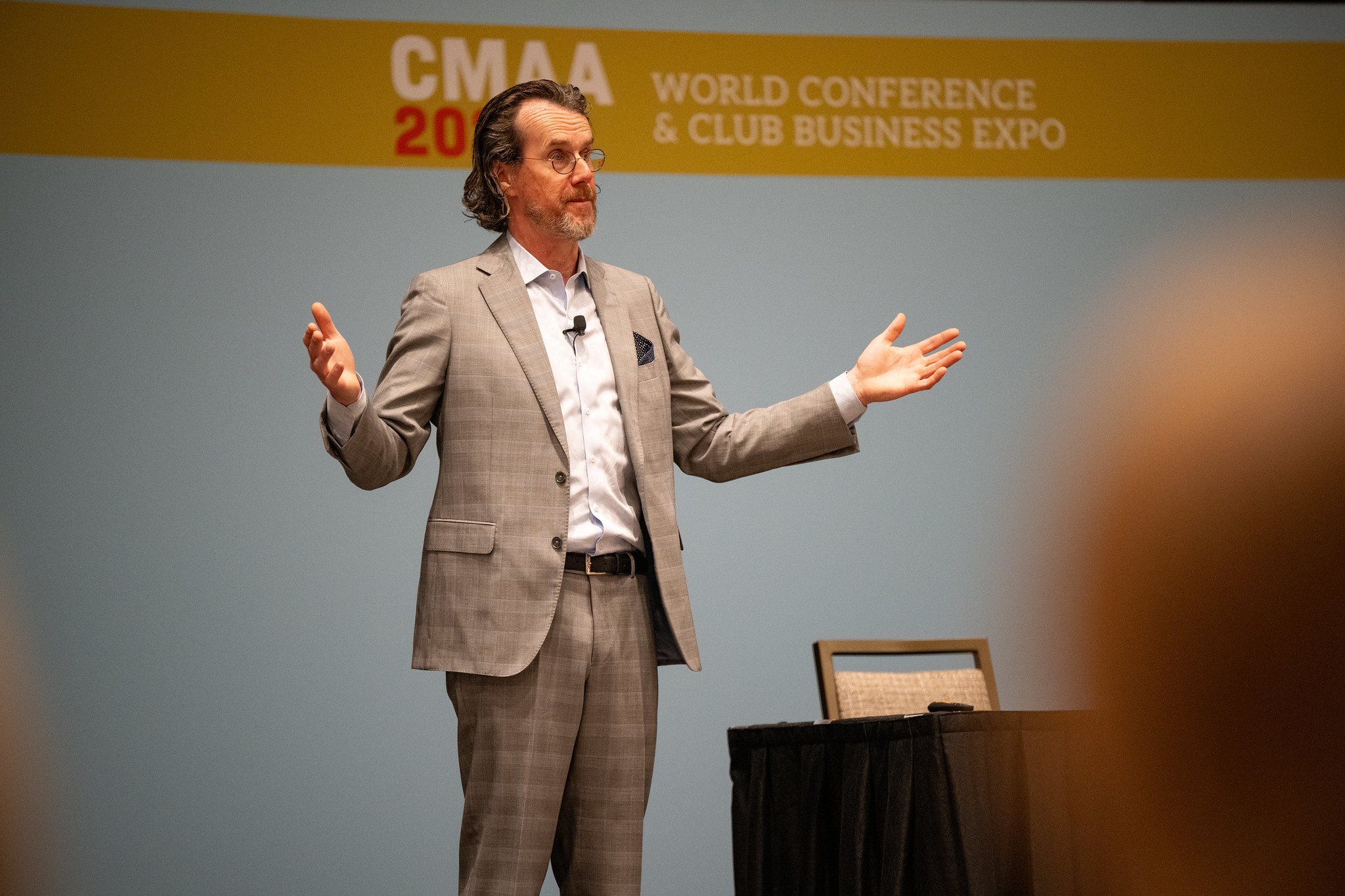 A man in a gray suit speaks at a Speaking Engagement during the CMAA World Conference & Club Business Expo, gesturing with both arms outstretched.