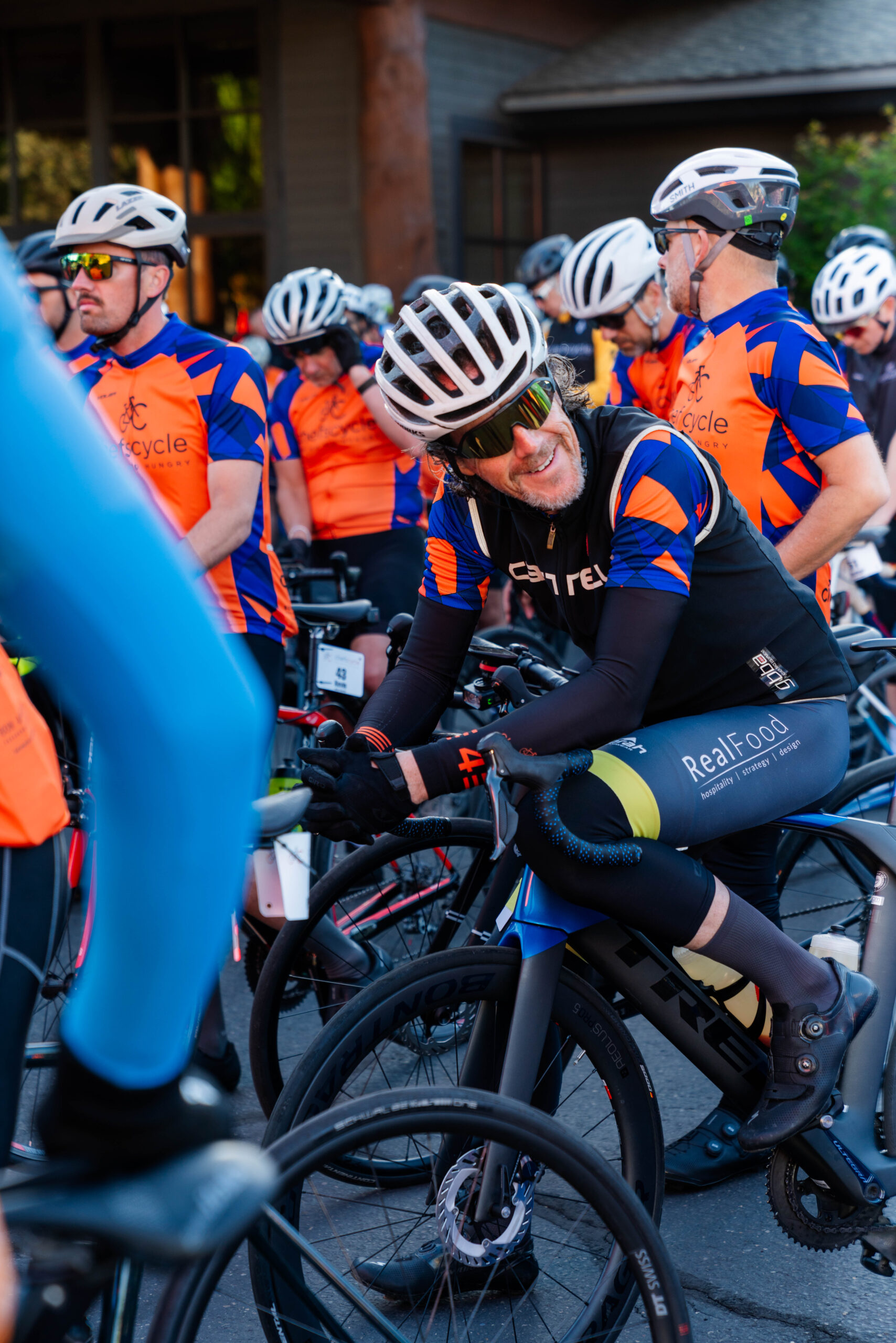 Cyclists in colorful jerseys gather at the starting line of a race or event, preparing with bikes and helmets.