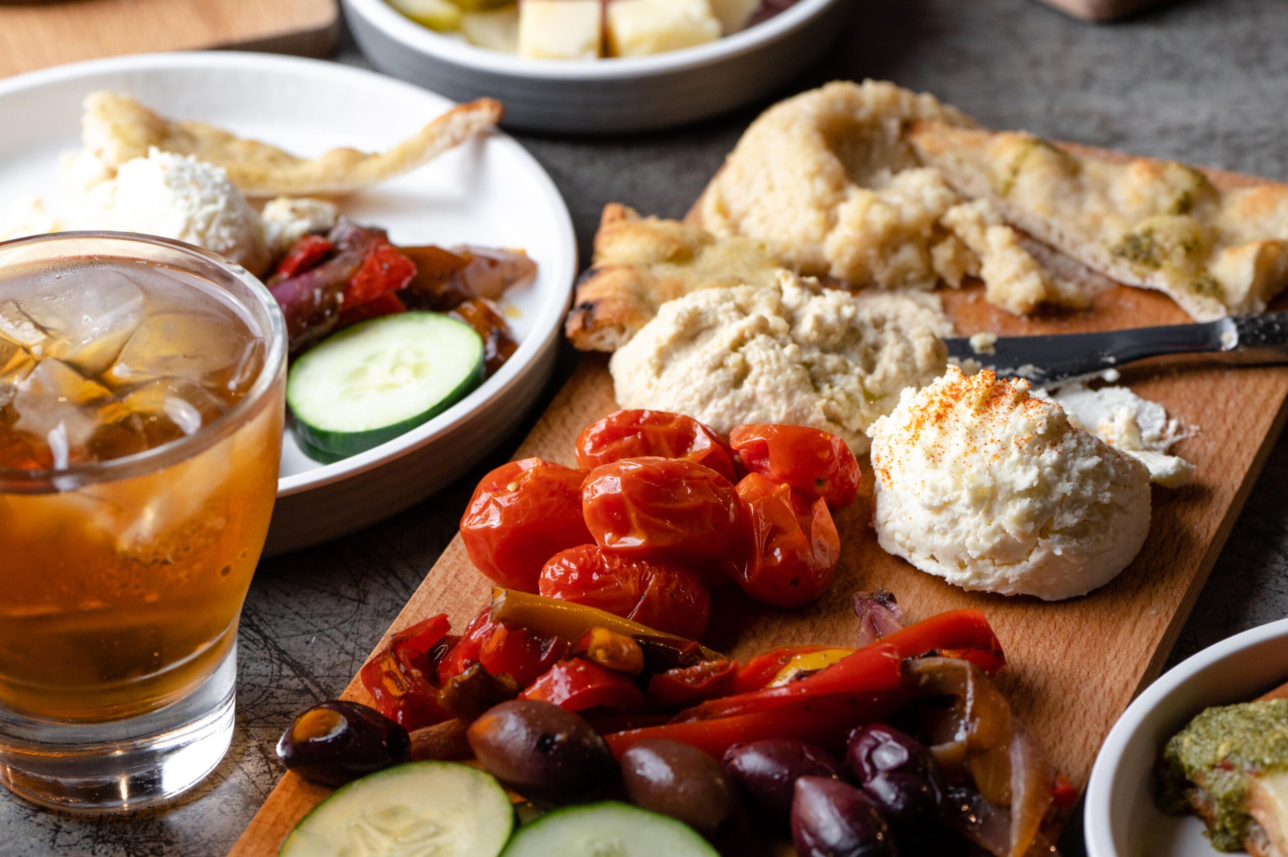 A wooden board with hummus, ricotta, roasted tomatoes, olives, cucumbers, and flatbread—delicious resources for any gathering—sits beside a glass of iced drink and a plate with cheese and vegetables.