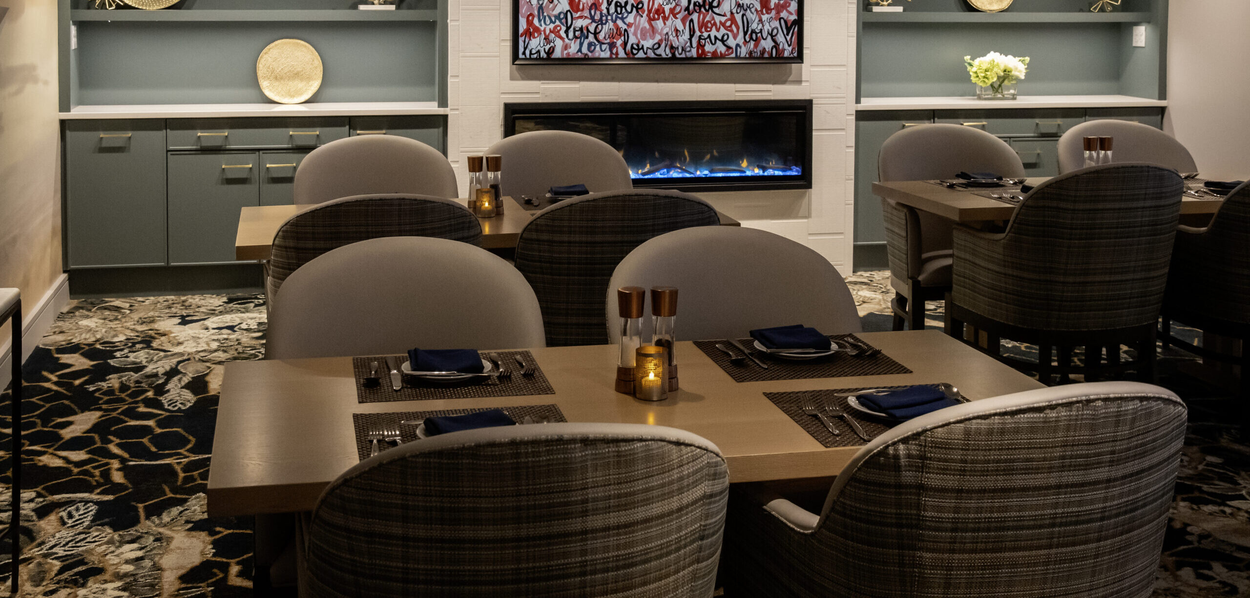Modern dining room with round tables, upholstered chairs, a fireplace, wall shelves displaying decorative plates, and a framed 