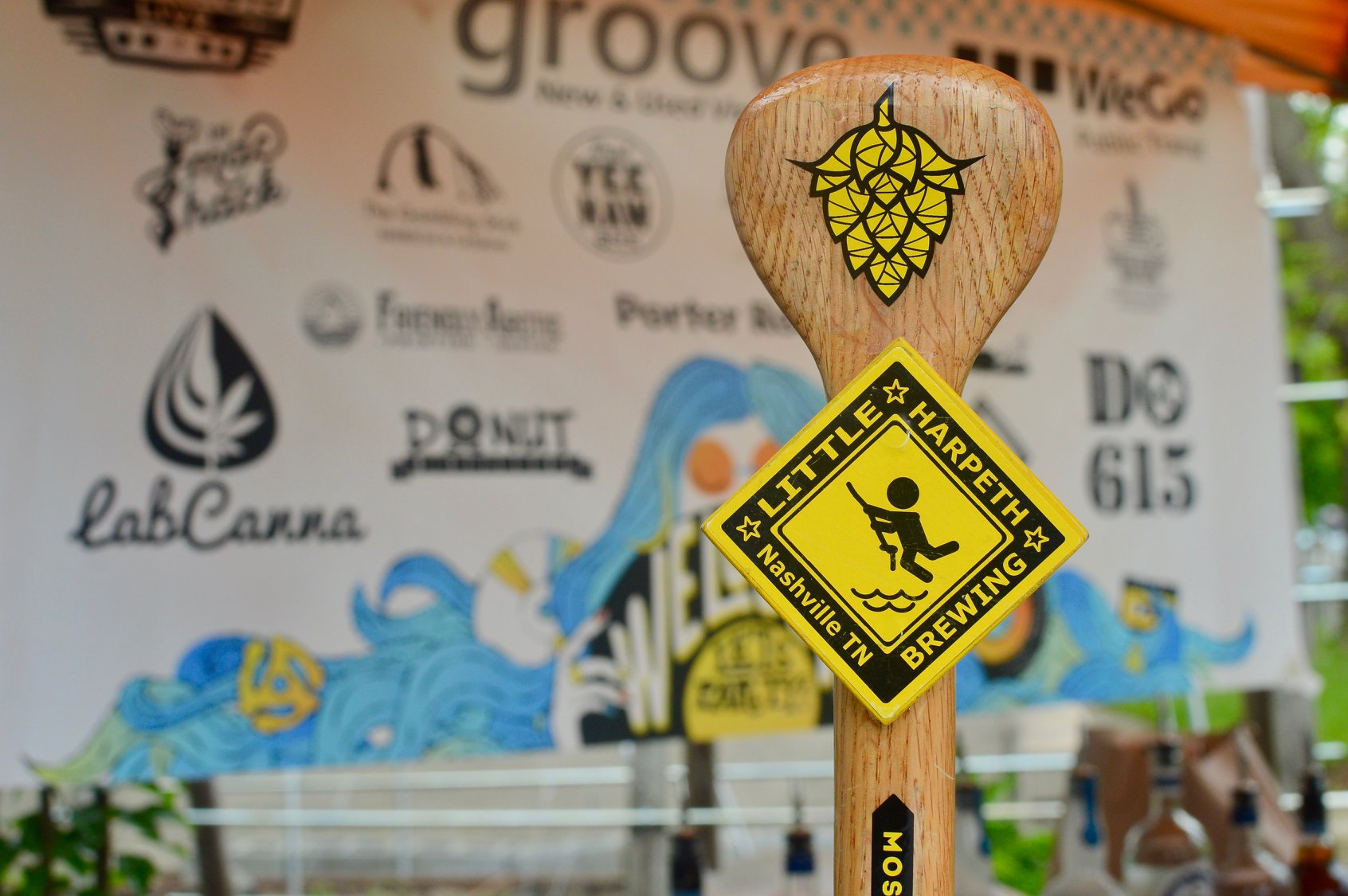 Close-up of a wooden beer tap handle displaying a Little Harpeth Brewing sticker, set against a backdrop featuring various brewery, business, and Procurement Services logos.